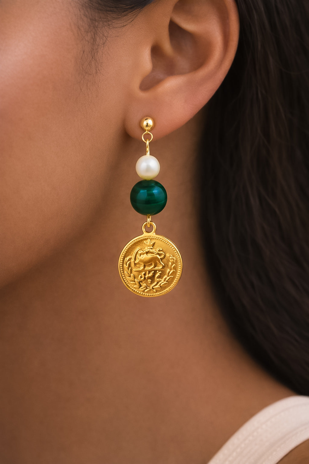 Close-up of an earring with a gold coin, pearl, and green stone on a person's ear.