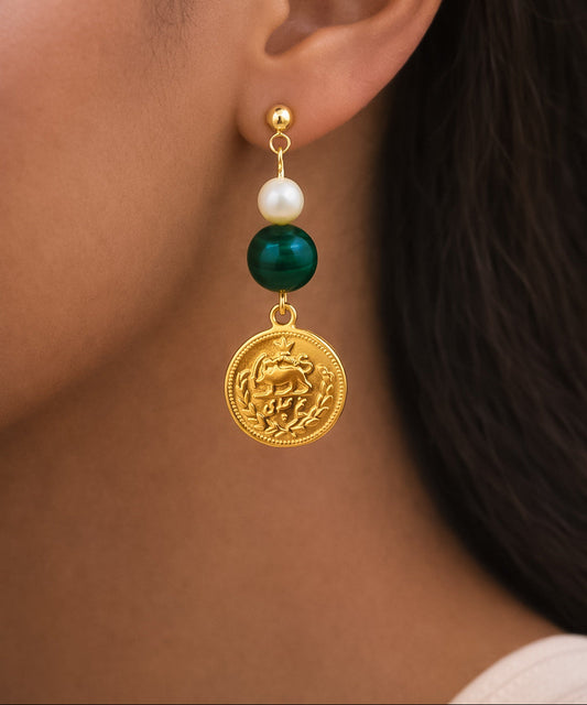 Close-up of an earring with a gold coin, pearl, and green stone on a person's ear.