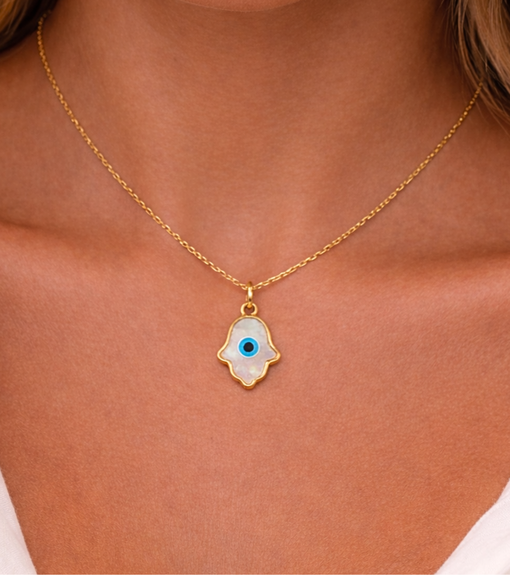 Gold necklace with a hamsa hand pendant featuring an eye design on a person wearing a white top.