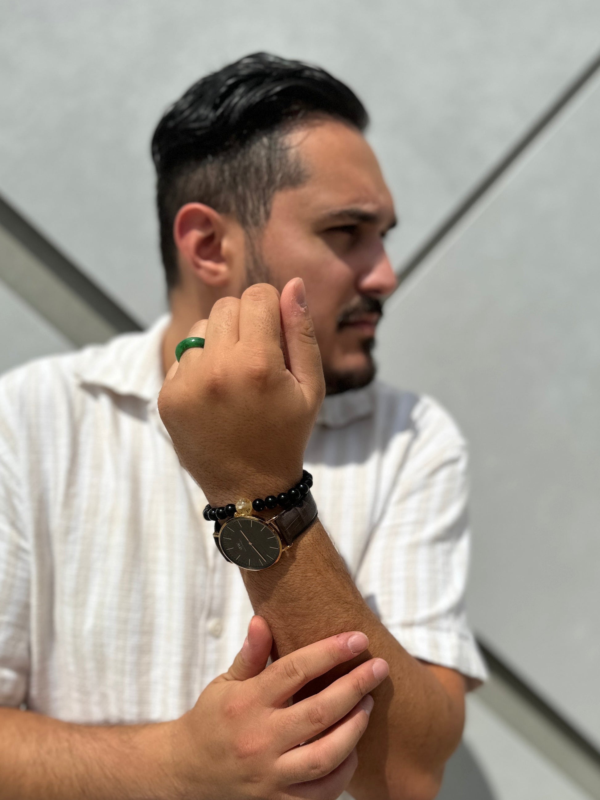 Man wearing a white shirt and multiple bracelets on his wrist, standing against a light-colored wall.