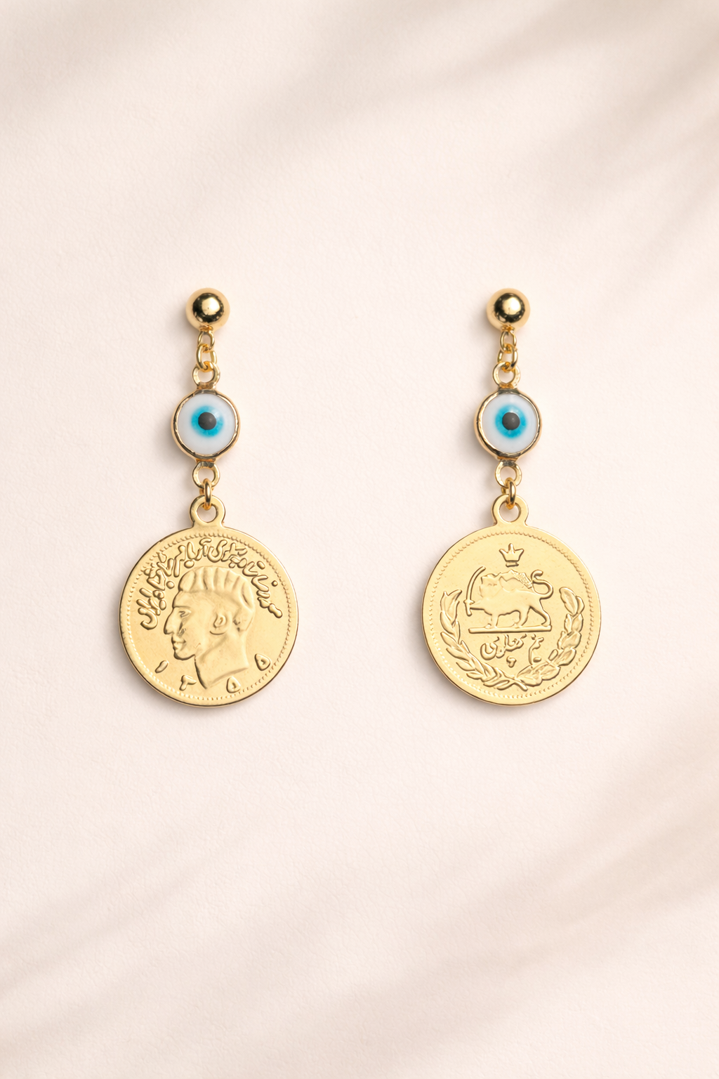 Pair of gold earrings with iranian coin designs on a light pink background